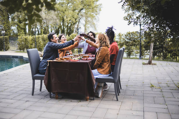 multiethnic group of friends sitting on a table by the pool celebrating a friendship toasting together - men and women best friends toasting arms up rising wine glasses outdoors - swimming pool water people sitting imagens e fotografias de stock