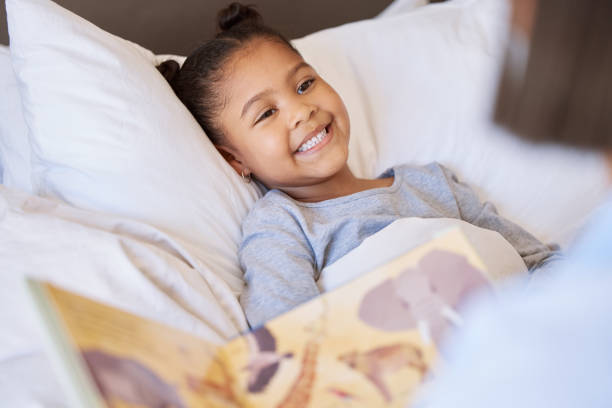 mother reading story book to daughter at bedtime. adorable little girl lying comfortable in bed and enjoying a story while being tucked in by mom. learning about animals - sleeping child bedtime little girls imagens e fotografias de stock