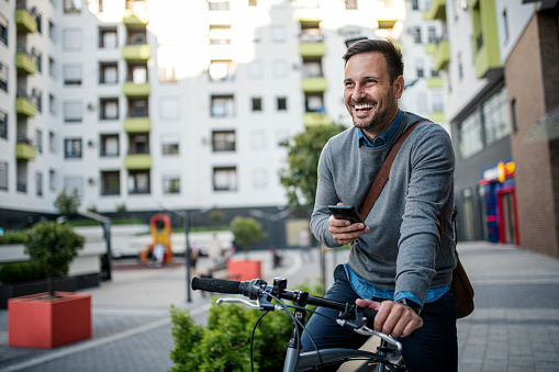 A college age student rides his bike through an urban setting on the way to school or work, wearing a backpack and helmet.  He checks his map on his smartphone to make sure he is going the correct direction.