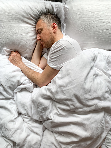 Middle aged adult man sleeping at home Cozy bedroom bed vibes white bedsheets Alone Early morning. White on white. Concept of comfort, relaxation.