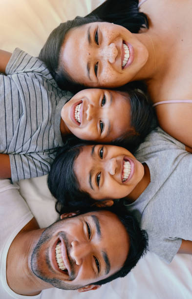 portrait d’une joyeuse famille métisse de quatre personnes en pyjama, allongée confortablement ensemble dans une rangée sur un lit souriant et prenant des selfies amusants à la maison. parents aimants avec deux enfants. des filles adorables qui crée - sweet tooth in a row photos et images de collection