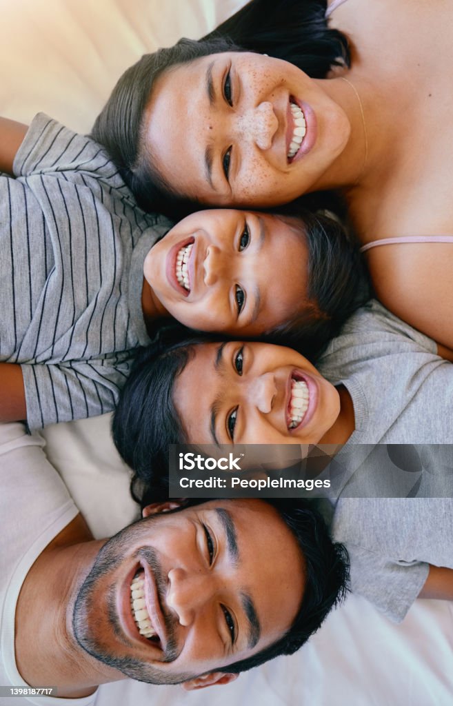 Portrait of a happy mixed race family of four in pyjamas lying cosy together in a row on a bed smiling and taking fun selfies at home. Loving parents with two kids. Adorable girls bonding with parents Family Stock Photo