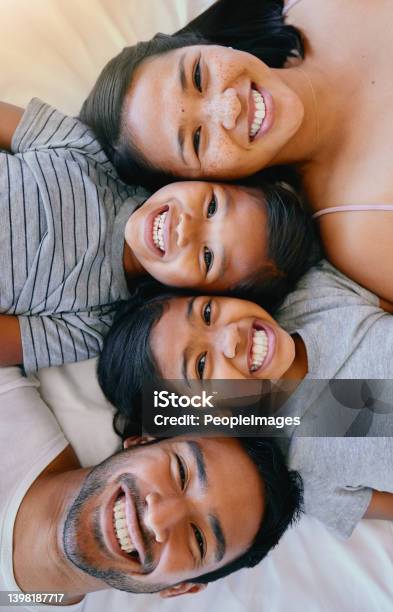 Portrait Of A Happy Mixed Race Family Of Four In Pyjamas Lying Cosy Together In A Row On A Bed Smiling And Taking Fun Selfies At Home Loving Parents With Two Kids Adorable Girls Bonding With Parents Stock Photo - Download Image Now