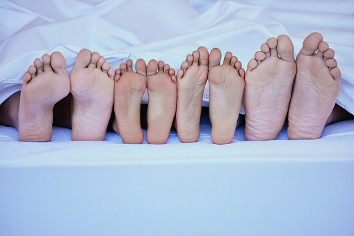 Mixed race closeup shot of family lying in bed showing their feet only under a clean white blanket and resting on a big comfortable bed. Unknown family relaxing and being lazy against bright copyspace