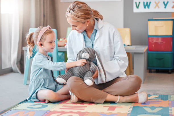 young caucasian paediatrician helping a little girl listen to the heartbeat of a toy with a stethoscope, small, happy child visiting the doctor for a checkup, playing on the floor together. - medical exam doctor patient adult imagens e fotografias de stock