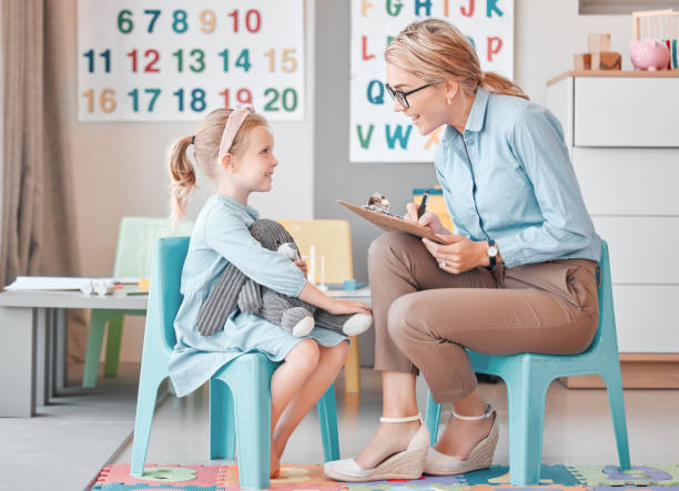 Young smiling caucasian child psychologist sitting with an adorable little girl in a clinic and using a clipboard for a consult. Cute girl talking to a mental health professional. Dyslexia and anxiety Young smiling caucasian child psychologist sitting with an adorable little girl in a clinic and using a clipboard for a consult. Cute girl talking to a mental health professional. Dyslexia and anxiety mental health professional stock pictures, royalty-free photos & images