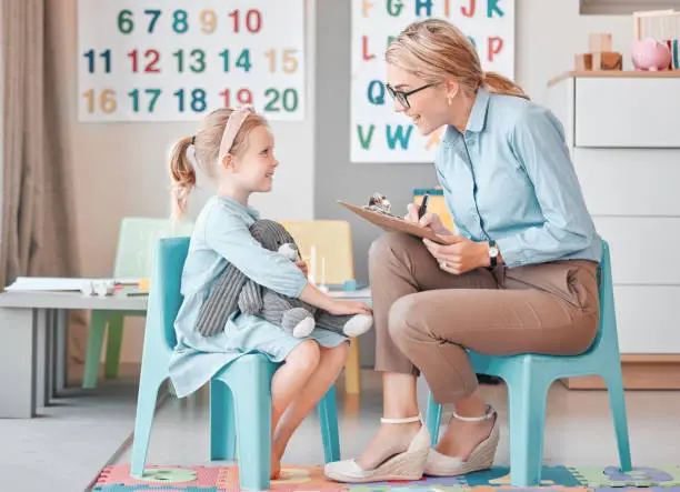 Photo of Young smiling caucasian child psychologist sitting with an adorable little girl in a clinic and using a clipboard for a consult. Cute girl talking to a mental health professional. Dyslexia and anxiety