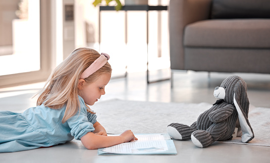 Little girl reading storybook at home with teddybear. Adorable caucasian girl lying on floor and reading a story to her stuffed animal