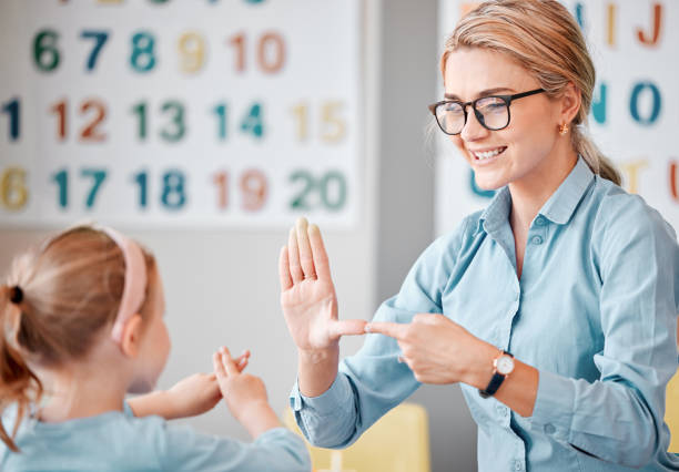 lächelnde kaukasische lehrerin mit brille, die mit gehörlosen schülerinnen mit handzeichensprache in einem klassenzimmer in der schule kommuniziert. gebärdensprachlehrerin in einer nachhilfeklasse für ein süßes weibliches kind mit hörbehinderung - deaf stock-fotos und bilder