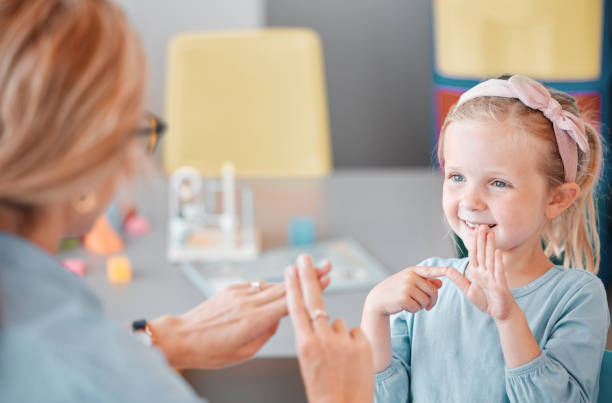 urocza uśmiechnięta mała kaukaska dziewczynka stojąca i używająca języka migowego do komunikowania się z psychologiem dziecięcym w klinice. specjalista ds. zdrowia psychicznego uczący głuche dziecko mówić gestami rąk - sign zdjęcia i obrazy z banku zdjęć