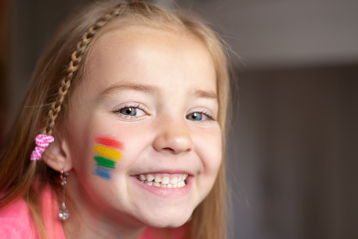 Close-up of a little girl with a rainbow flag painted on her cheeks. Portrait of a child from an LGBTQ family . Pride Day, LGBT family, concepts of rights and equality