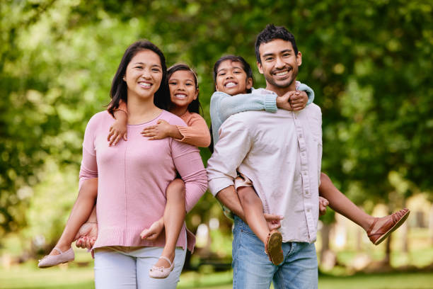 portrait of happy asian family standing close together in a park. adorable little girls enjoying free time with their mother and father on a weekend outside. smiling mixed race couple embracing their daughters - multi ethnic group family child standing imagens e fotografias de stock
