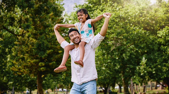 Happy mixed race father and cute playful daughter having fun at the park outdoors with copyspace. Carefree man carrying cheerful girl on shoulders for piggyback ride. Parent bonding with kid