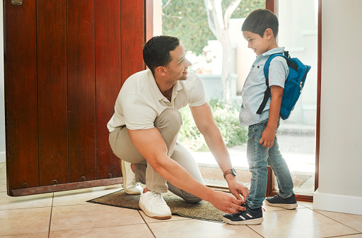 Happy single father tying his son's shoelaces before school in the morning. Adorable mixed race little boy carrying a backpack and getting help from his single parent. Hispanic man and child at home