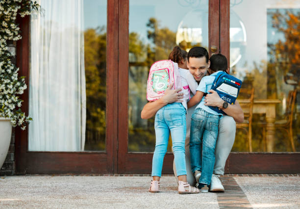 bambini che tornano da scuola e abbracciano il padre al loro ritorno. papà ha perso la piccola figlia e il figlio che tengono i bambini in braccio abbracciando la ragazza e il ragazzo mentre si trova vicino alla porta - visita foto e immagini stock