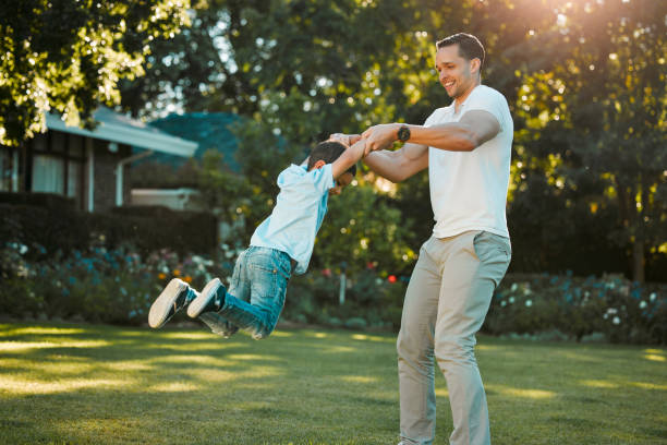 幸せなシングルファーザーは、庭で息子を外で回転させます。笑顔の混血の男と子供は、ゲームをしたり、裏庭で揺れたりしています。ひとり親は幼い男の子と絆を結び、空中で持ち上げる - swing child swinging front or back yard ストックフォトと画像