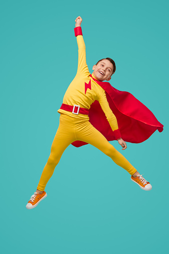 Carefree child in superhero costume jumping above ground with raised arm and celebrating success on blue background in studio