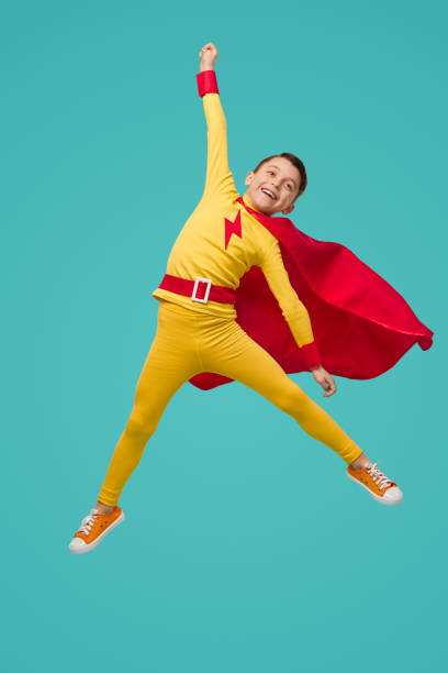 Excited superhero boy jumping and celebrating victory Carefree child in superhero costume jumping above ground with raised arm and celebrating success on blue background in studio jump jet stock pictures, royalty-free photos & images