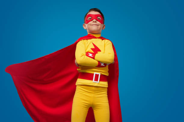 Low angle of proud self assured brave boy in bright yellow superhero outfit with flying red cape, and mask standing with arms crossed against blue background