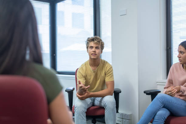 Teenage boy sharing life experience with peers in group therapy A teenage boy seeking counseling at his school talks while attending a group therapy session. teenagers only teenager multi ethnic group student stock pictures, royalty-free photos & images