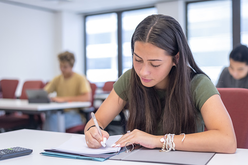 A female Eurasian student in high school writes in a notebook while in class at school.