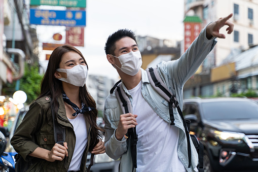 Asian romantic couple wear mask, travel in the city for honeymoon trip. Young new marriage man and woman backpacker tourist feel cheerful and excited, enjoy spending time on holiday vacation together.