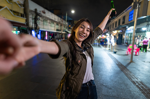 Portrait of Asian woman traveler hold boyfriend's hand, walk on street. Young man and woman tourist travel in city spend time on vacation together and drink bottle of beer at dark night on the road.