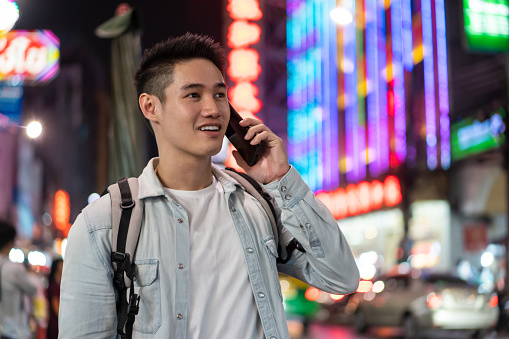 Asian attractive young man backpacker talking on phone walk in city. Handsome male tourist traveler travel alone on street, using smartphone while enjoy spend time on holiday vacation trip at night.