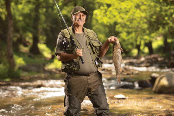 smiling mature fisherman holding a carp fish and standing in a river - anglerfish imagens e fotografias de stock