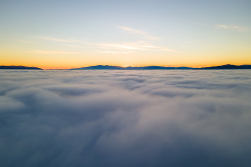 Aerial view of vibrant yellow sunrise over white dense clouds and distant mountains on horizon.