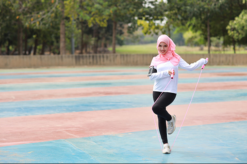 Athletic young asian woman in muslim sportswear standing and skipping rope outdoor for morning exercise. Active girl work out on biceps exercise with green tree background. Sport concept