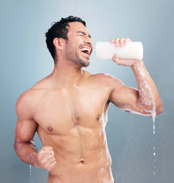 un jeune homme métis sexy et musclé chantant sous la douche sur un fond de studio bleu. un gars en forme qui s’amuse avec une bouteille de savon ou de shampoing comme un faux micro pendant sa routine quotidienne de soins corporels - male torso abdominal muscle the human body photos et images de collection