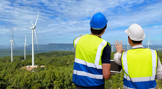 Windmill engineer man and worker standing and planing renewable energy technology or alternative ecology project for future. Electrician engineer looking and checking wind turbines site with computer