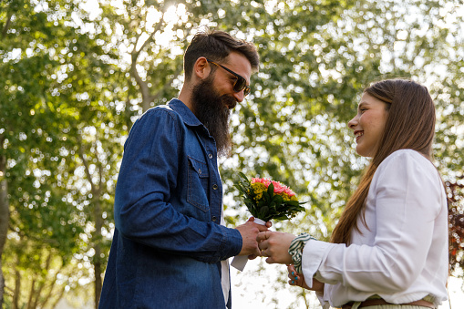 Low angle view of affectionate young man smiling at his loving girlfriend while giving her a bouquet of flowers under green trees at the park.