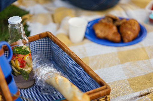 Selective focus shot of a humble picnic spread. A vintage picnic suitcase, glass bottle with water with pieces of mint, strawberry, cucumber and lemon in in, a plate with croissants and a baguette to be enjoyed.