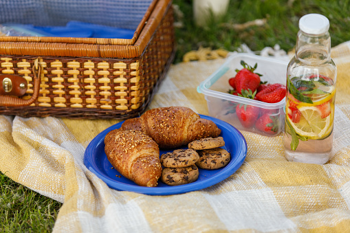 Flat lay of a vintage picnic suitcase, a glass bottle with water with pieces of fruit floating inside, a plate with croissants and chocolate chip cookies and bowl with fresh strawberries to be enjoyed during picnic.