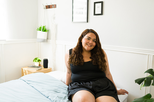 Cheerful big young woman sitting in bed. Hispanic obese woman enjoying a leisure day in her beautiful bedroom