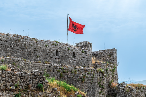 Shkoder, Albania - June 21, 2021: The Albanian flag flies over the ruins of Rozafa Castle against a blue sky on a summer day, Albania. Ancient fortification stone walls with arched holes, close-up
