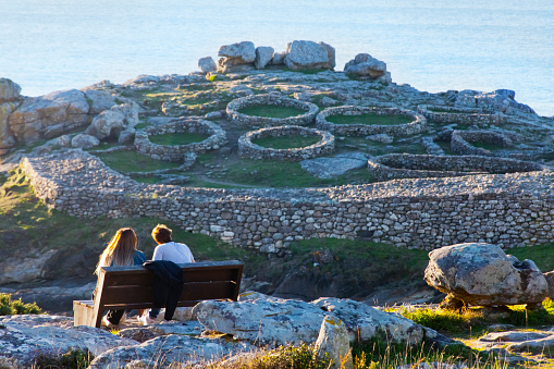 Porto do Son, Spain - January 12, 2022: Rear view of  young couple sitting on a bench watching Atlantic ocean and Hill fort 'Castro' of Baroña, A Coruña  province, Galicia, Spain. Headland in front of the Atlantic ocean set 1st century BC. The Hill fort 'Castro' of Baroña is completely public free access.