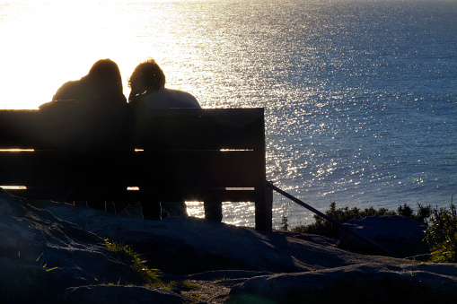 Rear back lit view of  unrecognizable young couple sitting on a bench watching sunset over Atlantic ocean, Porto do Son, A Coruña  province, Galicia, Spain.