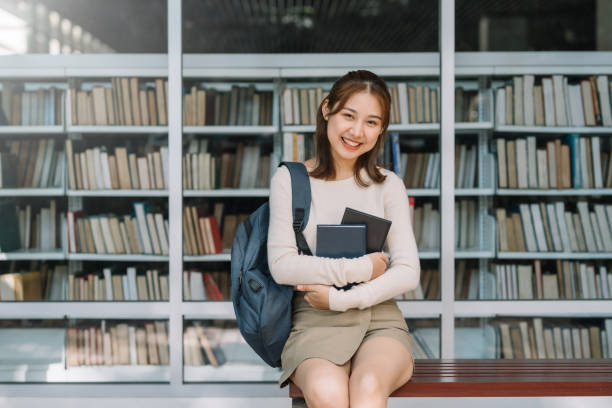 Young Asian female student sitting holding book looking at camera. Library background stock photo