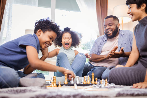 lustige afroamerikanische kinder, die mit mama und papa zu hause schach spielen - togetherness learning playful mother stock-fotos und bilder