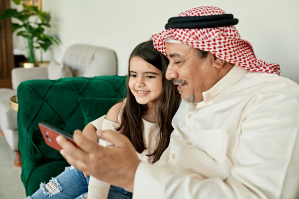Mature Saudi man and granddaughter taking selfie at home Middle Eastern man in traditional attire with arm around child in casual western clothing,  sitting together on sofa and smiling at smart phone. kaffiyeh stock pictures, royalty-free photos & images