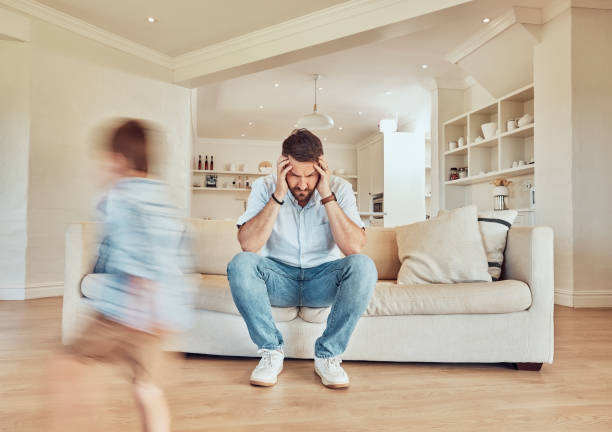 istock Young caucasian father suffering from a headache while his son runs in the lounge at home. Carefree boy running in the living room while his dad looks tired. Man looking upset while his child plays 1398246850
