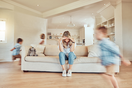 Stressed mother sitting on the sofa while her adhd children run around the living room. Hyperactive son and daughter giving single parent a headache. Active siblings chasing each other in the house