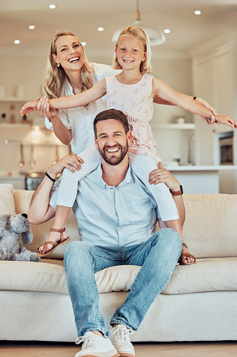 Portrait of excited little playful girl sitting on father's shoulders and holding mother's hands while pretending to fly at home. Happy caucasian parents enjoying fun quality time with daughter