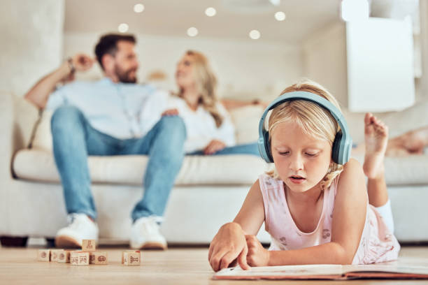 mała kaukaska dziewczynka czytająca książkę z opowiadaniami, w słuchawkach słuchająca muzyki z rodzicami w tle. dziecko czyta bajkę i słucha muzyki z mamą i tatą w domu - six animals audio zdjęcia i obrazy z banku zdjęć