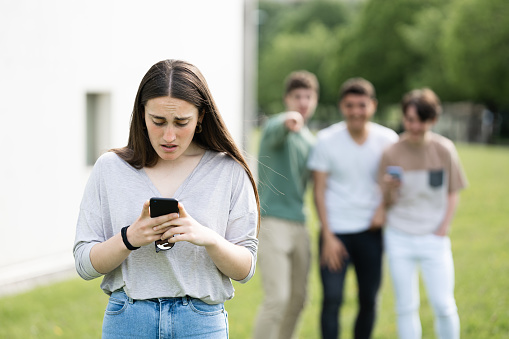 Surprised teenager girl looking at phone while friens are laughing at her. Cyber bullying concept.