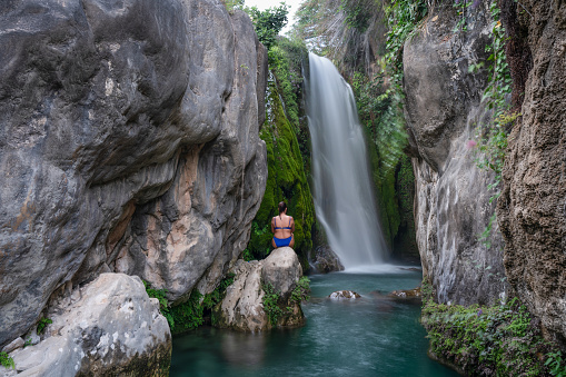 Back view of a pensive woman wearing a blue swimwear sitting on rock in front of a silky waterfall