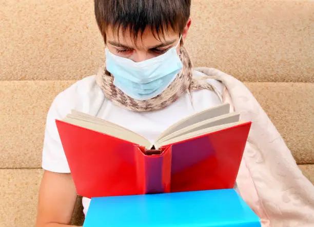 Sick Teenager in Flu Mask read a Book on the Sofa at the Home